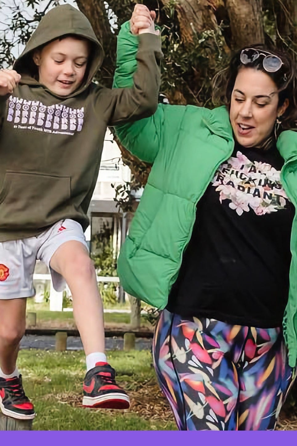 A mum and her young son laughing as they jump hand in hand in a park