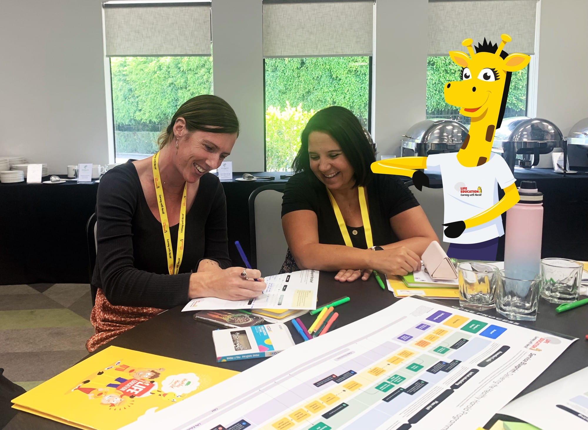 Two teachers smiling over service blueprint worksheets at a Life Education workshop, with an illustrated Harold the giraffe standing alongside