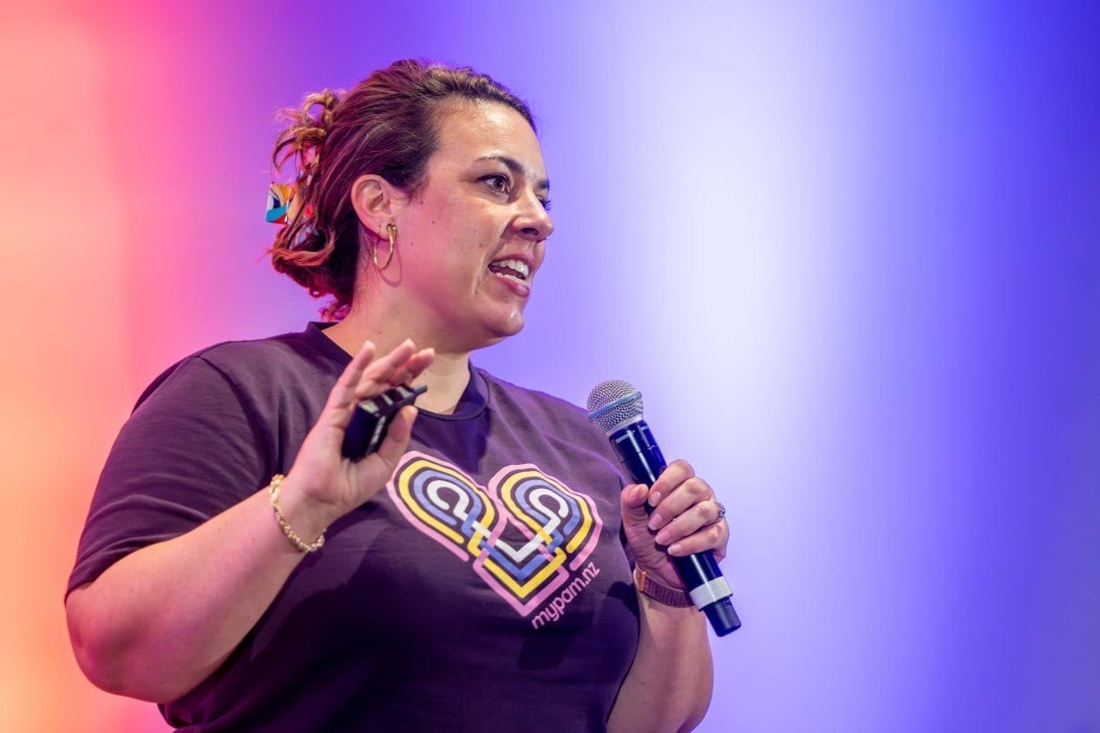 PAM founder Nicole Retter speaking on stage, microphone in hand, wearing a mypam.nz t-shirt