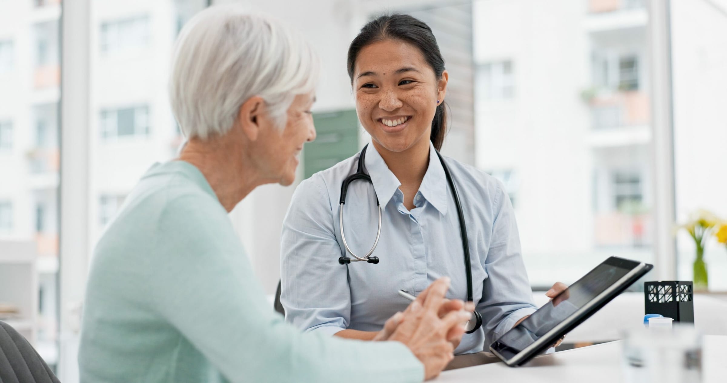 Nurse with a stethoscope holding a tablet while talking with an older patient during a consultation