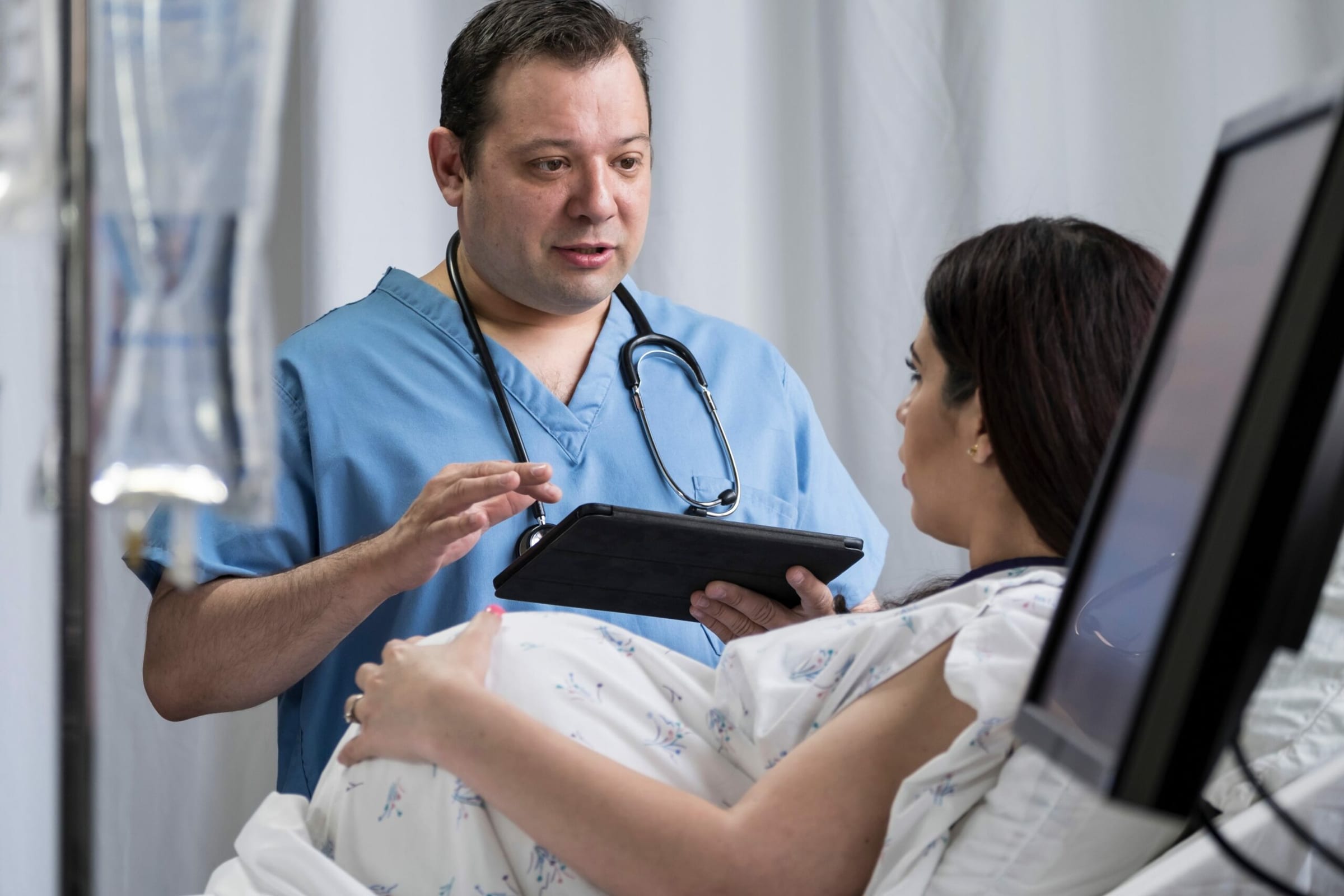 Male nurse in scrubs holding a tablet while talking with a patient in a hospital bed