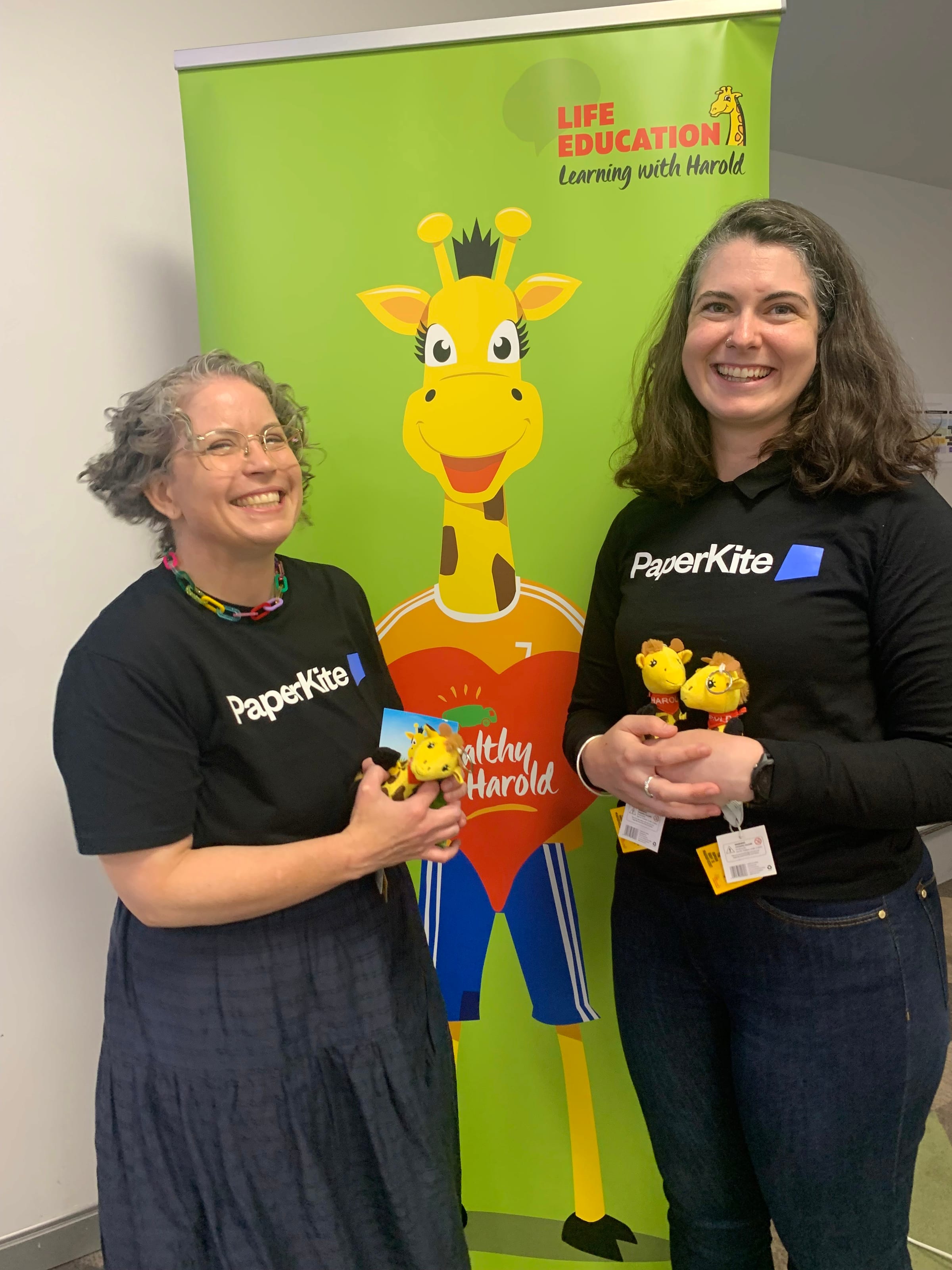 Two PaperKite team members holding Harold the giraffe plush toys in front of a Life Education banner