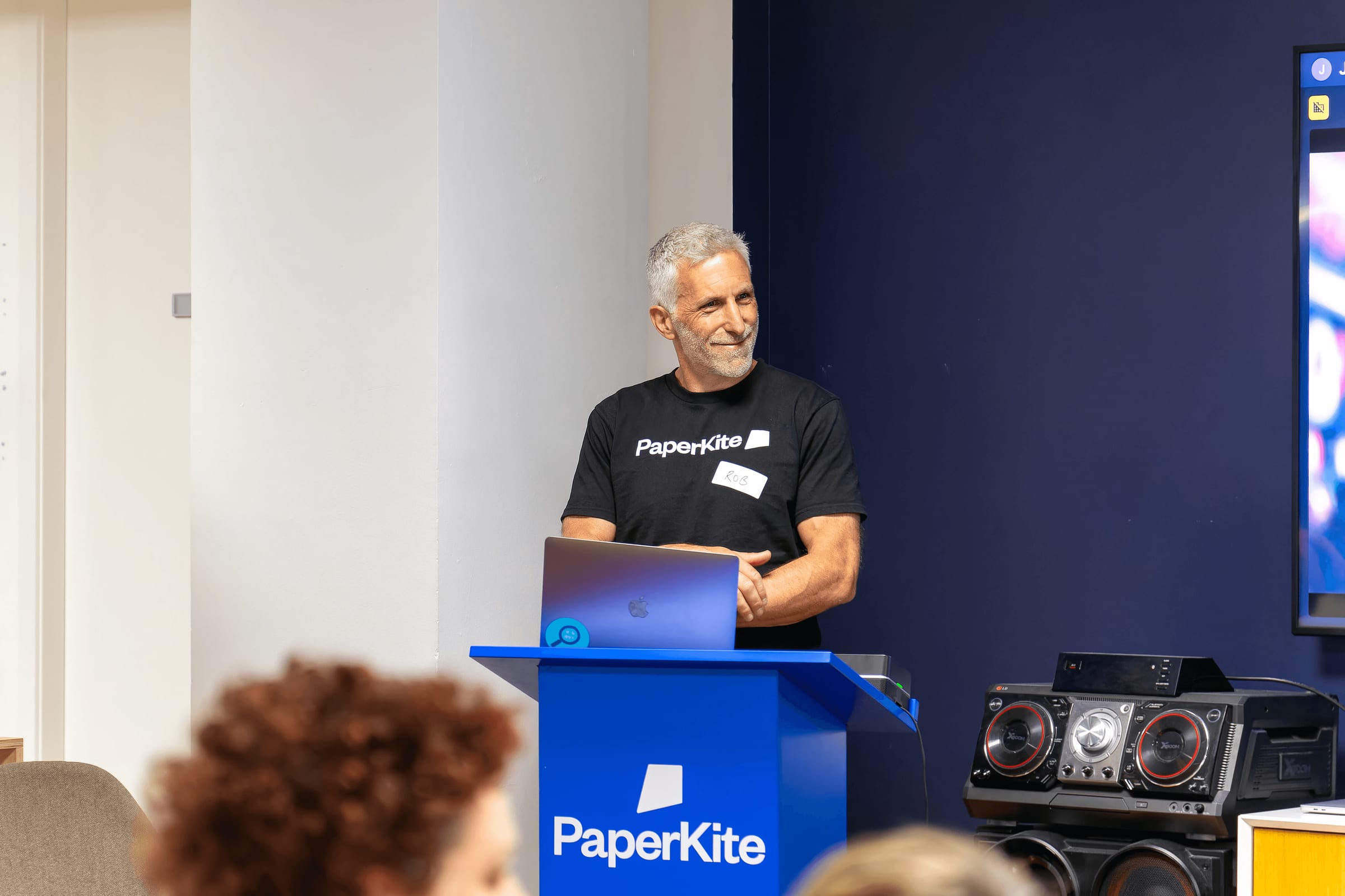A PaperKiter standing at a blue podium branded with the PaperKite logo, preparing to deliver a presentation